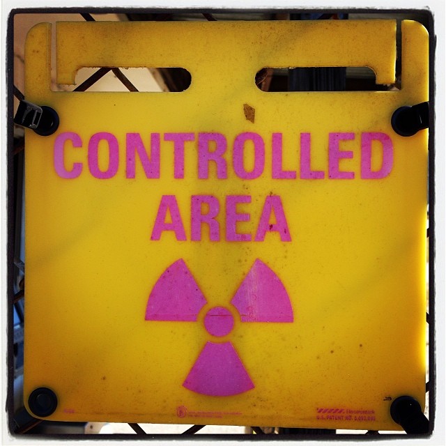 build connection and engagement: photograph of a square yellow plastic sign clamped to a wire mesh fence. The pink sign says "CONTROLLED AREA" with a radioactivity symbol underneath.