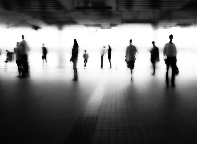 life story exercise: A blurred black-and-white photograph of a group of people standing apart from each other in a large hall.