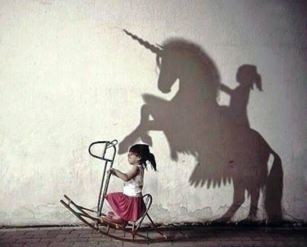 integrating your inner child: an image of a little girl rocking on a horse made of metal pipe. On the white wall behind her is the shadow of her riding a much larger, rearing unicorn